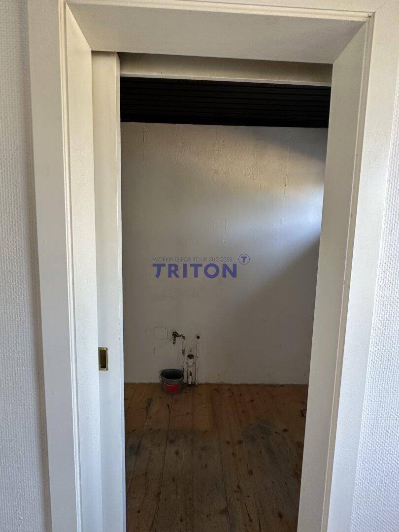 Blick durch eine Tür in einen kleinen Raum mit Holzfußboden und Wasseranschlüssen.