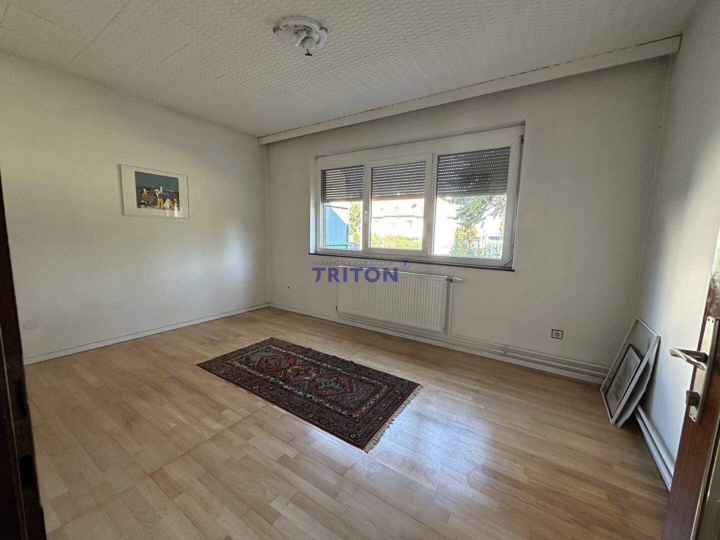 Heller Raum mit Holzfußboden, strukturierter Decke und Fenster mit Jalousien.