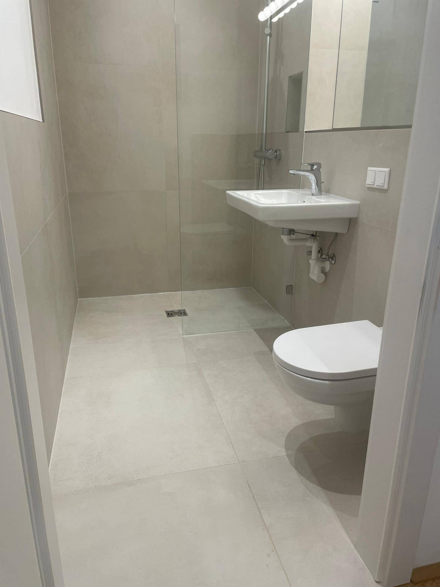 Modernes Badezimmer mit hellgrauen Fliesen, ebenerdiger Dusche und wandhängendem WC.