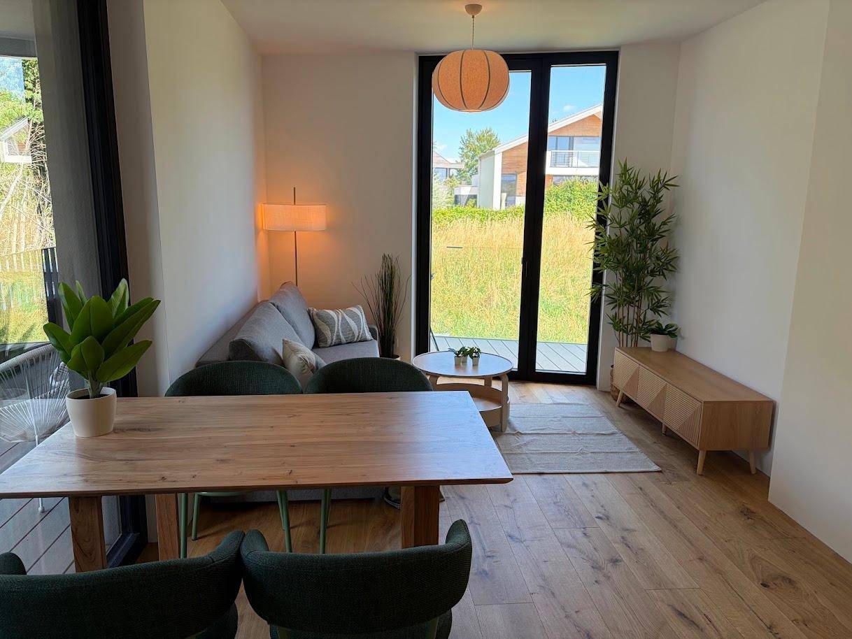 Heller Wohn-Essbereich mit Holzboden, Esstisch, Sofa und großen Fenstern mit Blick ins Grüne.