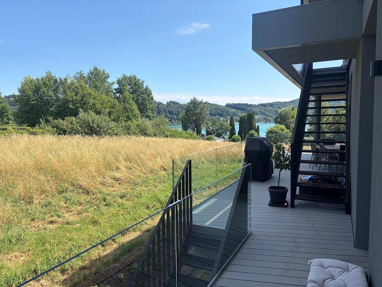 Geräumige Terrasse mit Glasgeländer, Außentreppe und weitem Blick über grüne Felder zum See.