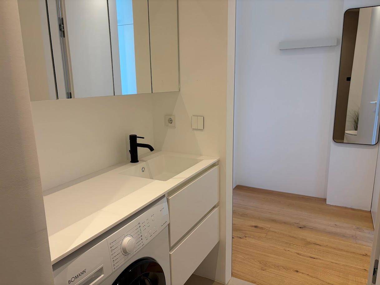 Funktionaler Waschraum mit Waschbecken, Spiegelschrank und integrierter Waschmaschine auf Holzboden.
