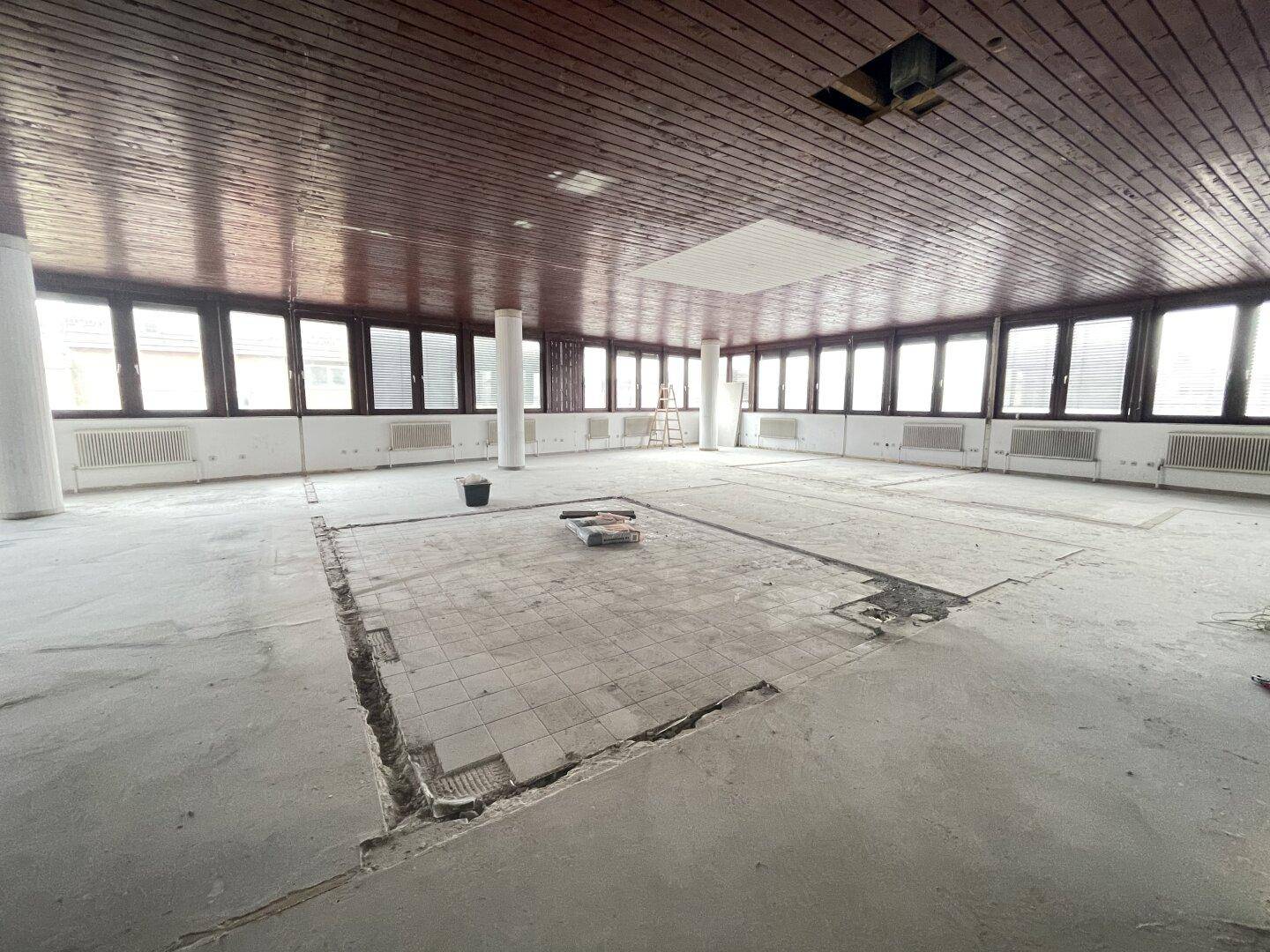 Großer, leerstehender Büroraum mit vielen Fenstern, teilweise entferntem Bodenbelag und einer Holzdecke.