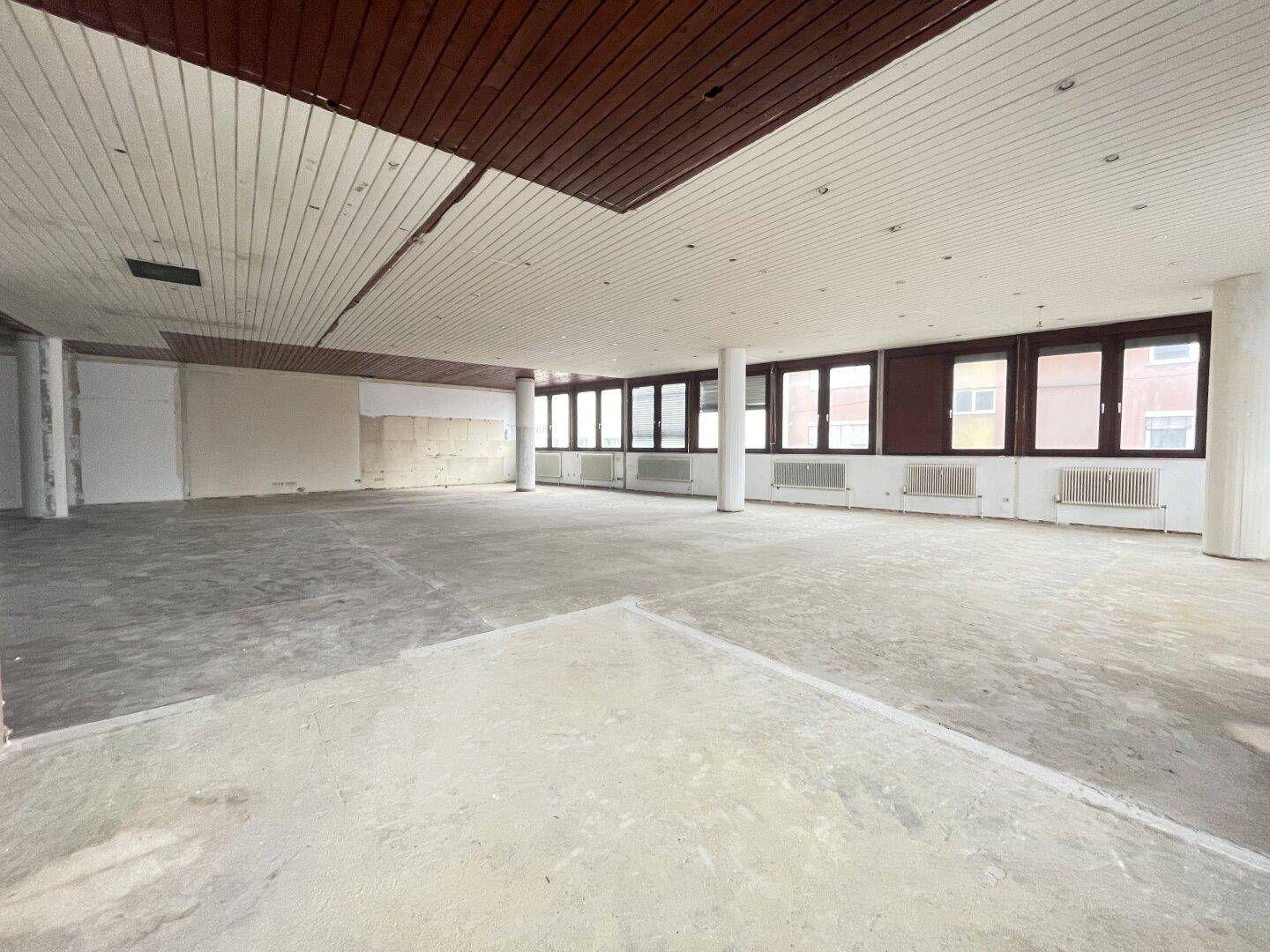 Weiterer Blick in den großen, leeren Büroraum mit Holz- und Paneeldecke sowie kahlen Wänden, bereit zur Renovierung.