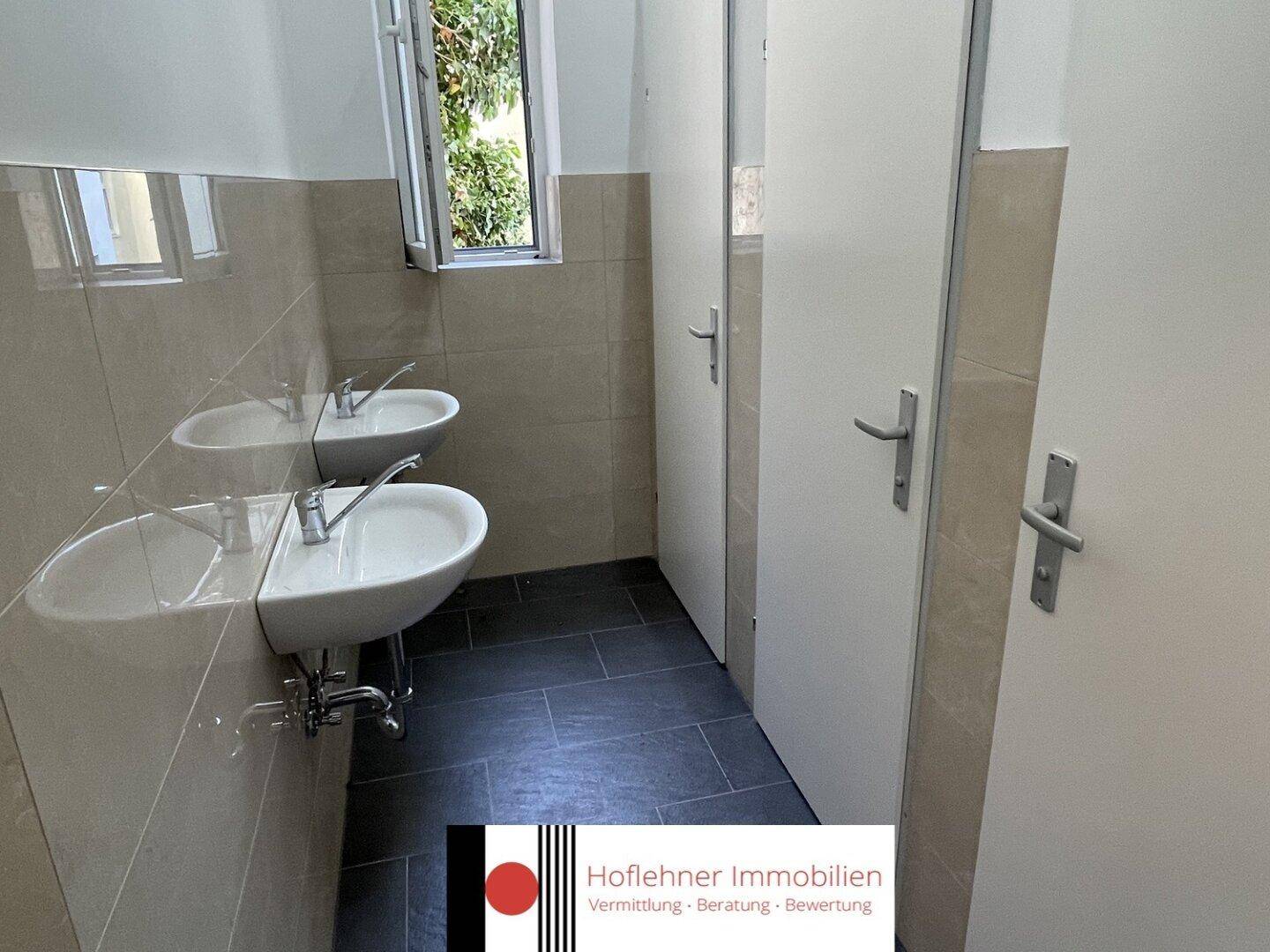 Moderner Sanitärbereich mit zwei Waschbecken und dunklen Bodenfliesen, inklusive Fenster für Tageslicht.