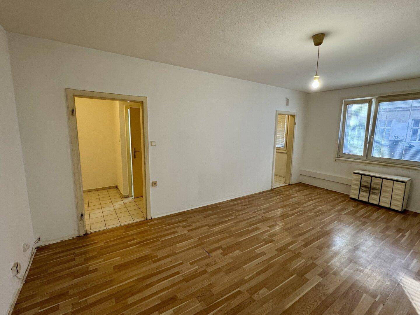 Heller Wohnraum mit Parkettboden, Fenster und Zugängen zu weiteren Zimmern der Wohnung.