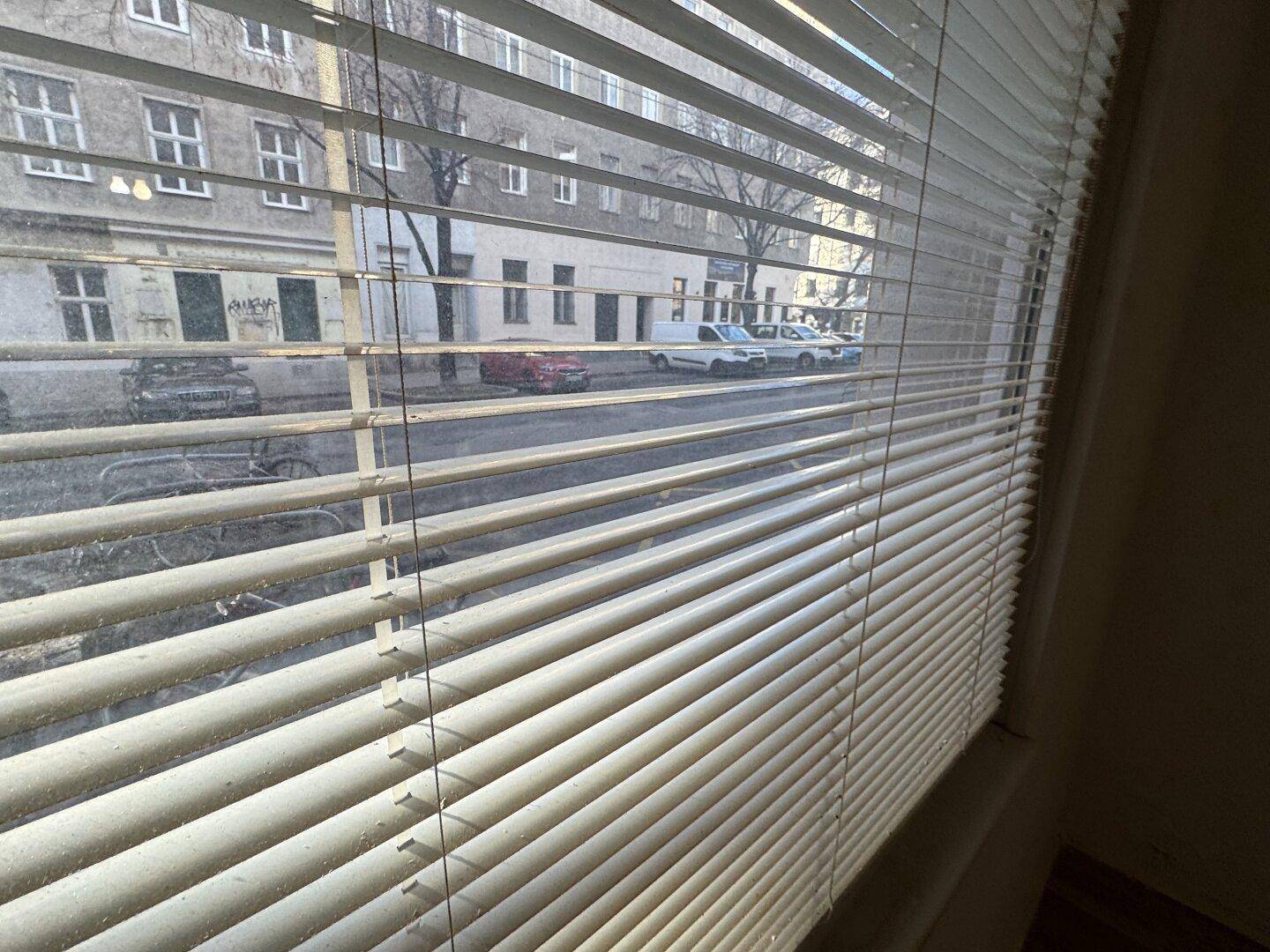 Blick aus dem Fenster durch Jalousien auf eine belebte Straße mit Gebäuden und parkenden Autos.