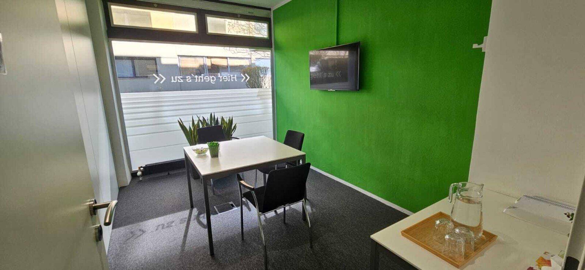 Besprechungsraum mit grün gestrichener Wand, Tisch, Stühlen und Fenster.