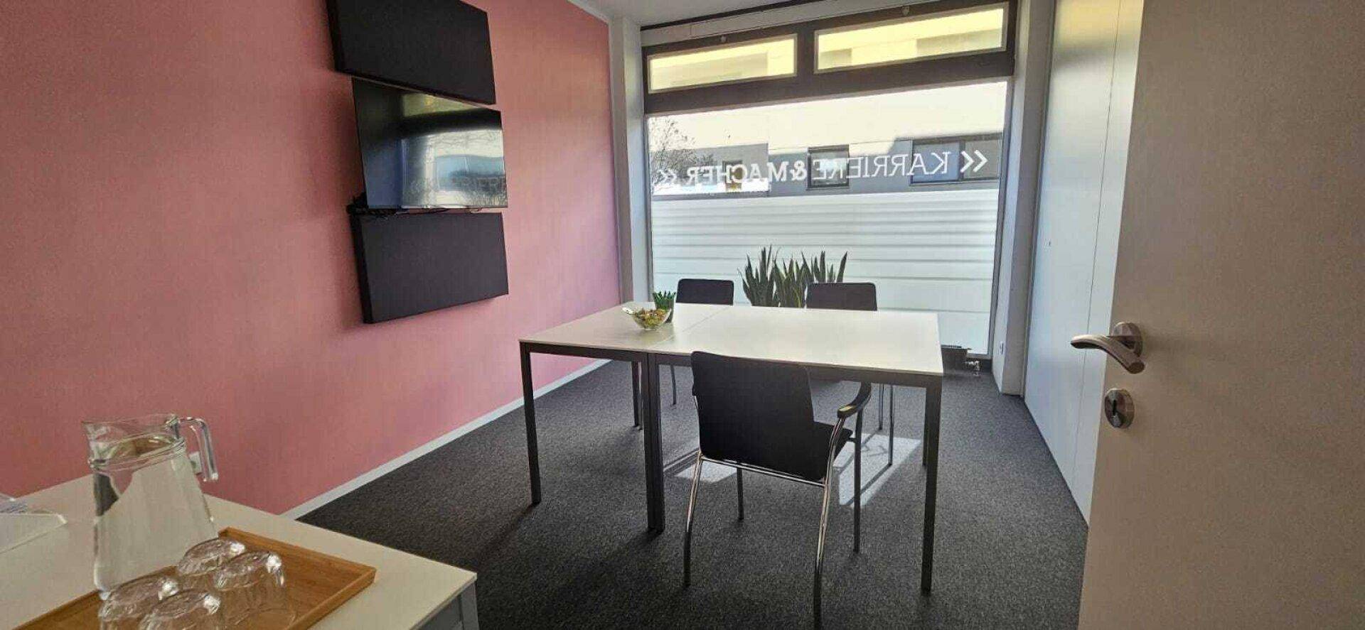 Besprechungsraum mit rosa Wand, Tisch, Stühlen und Fenster mit Blick nach draußen.