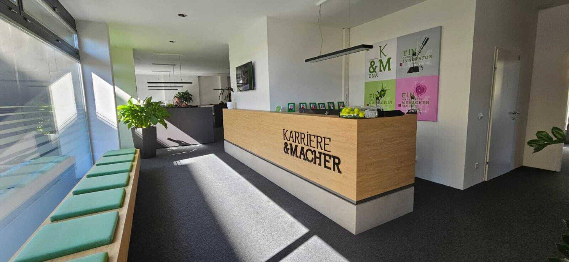 Moderner Empfangsbereich mit Holztheke, dunklem Teppichboden und großen Fenstern.