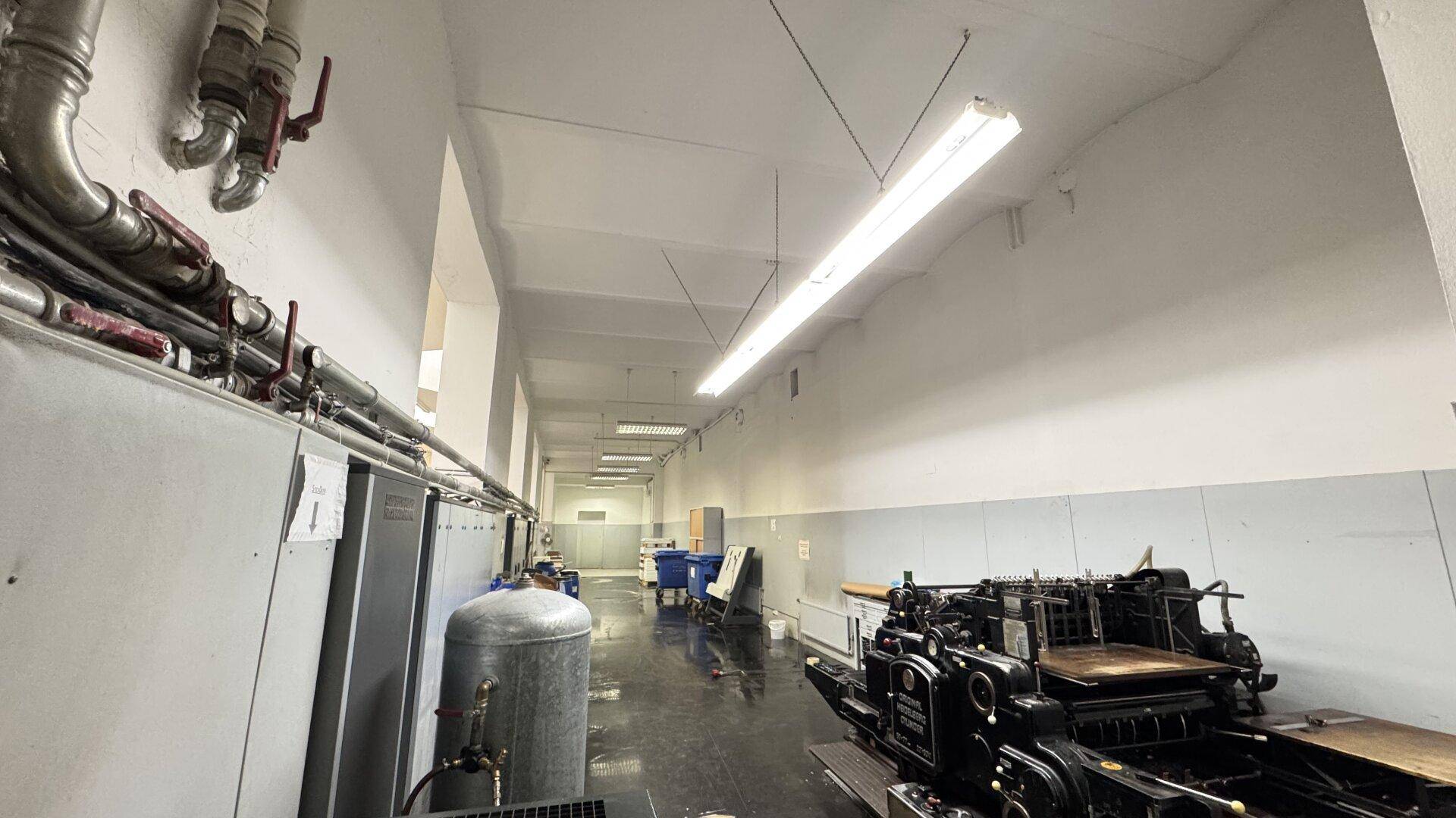 Industrieller Bereich mit sichtbaren Rohrleitungen, Maschinen und langen Leuchtstofflampen an der Decke.