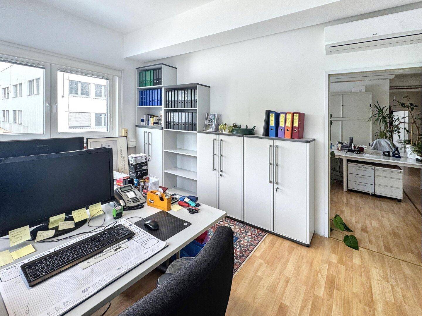 Heller Büroraum mit Schränken, einem Arbeitsplatz und Blick nach draußen durch die Fenster.