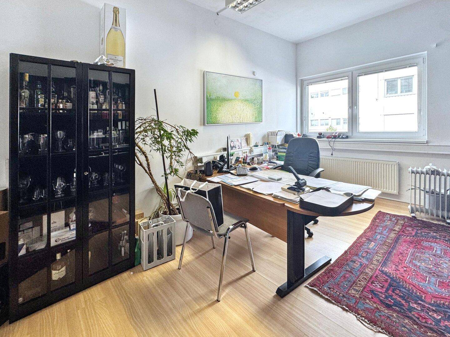 Büro mit einem Schreibtisch, einem Glasschrank und einem Heizkörper unter dem Fenster.