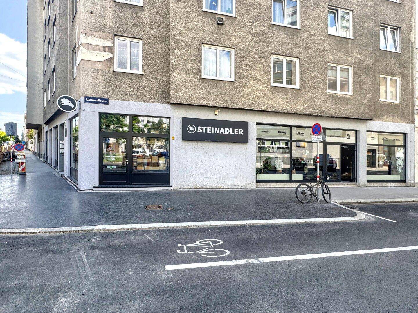 Gebäudeecke mit einem modernen Geschäftslokal im Erdgeschoss und einer Fahrradspur auf der Straße.