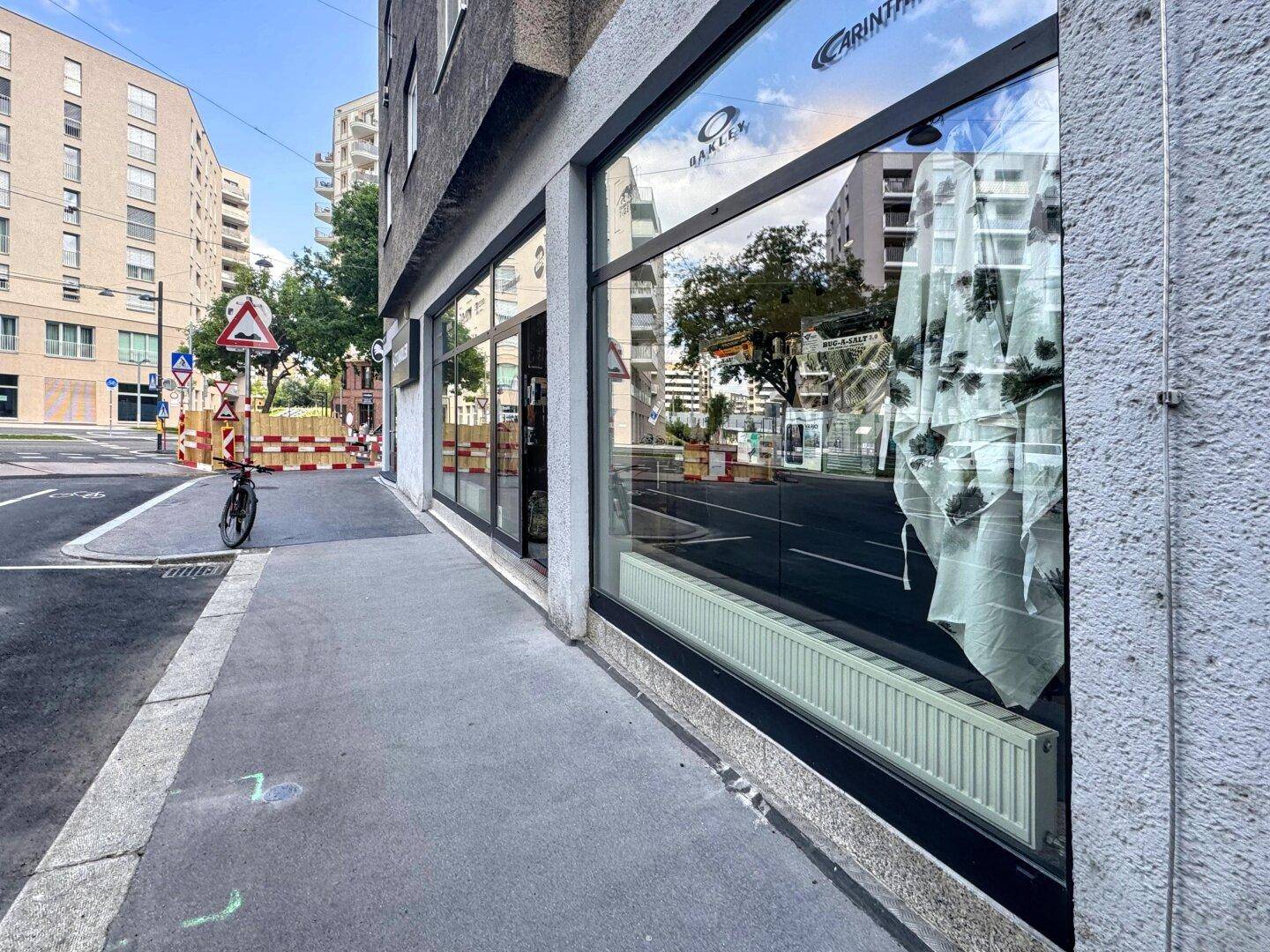 Blick entlang des Bürgersteigs mit großen Schaufenstern, die die städtische Umgebung reflektieren.