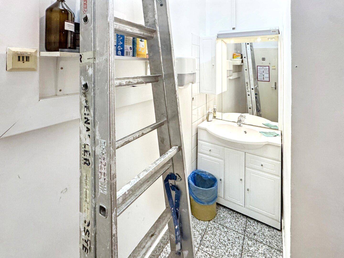Badezimmer mit Waschbecken, Spiegel und Unterschrank, derzeit mit einer Leiter für Wartungsarbeiten.