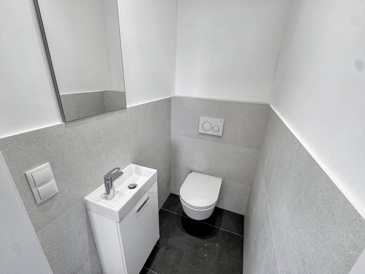Modernes Gäste-WC mit Wand-WC, kleinem Waschbecken und grauen Fliesen.