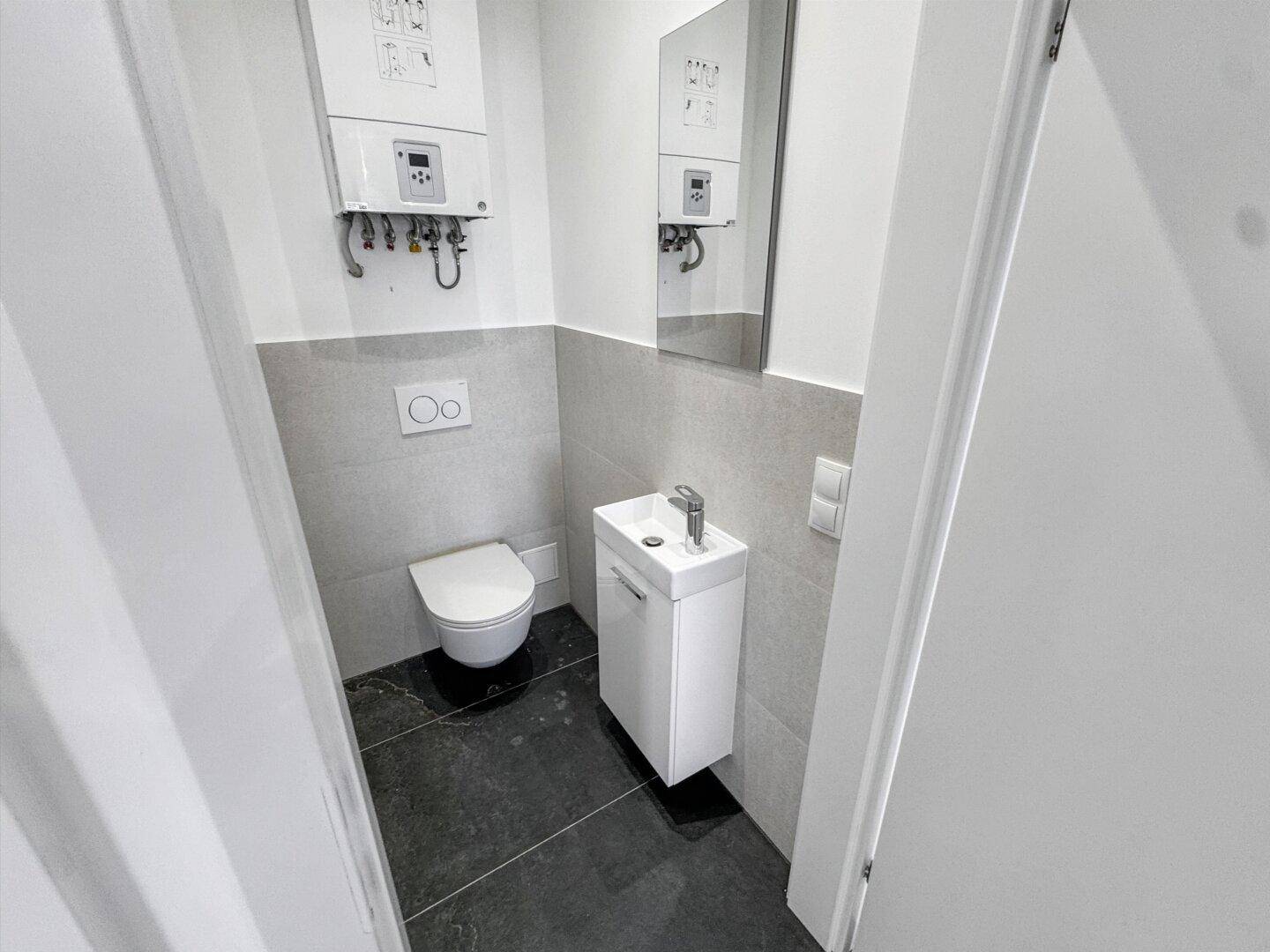 Kompaktes Gäste-WC mit Wand-WC, Waschbecken und Heiztherme, modern gefliest.
