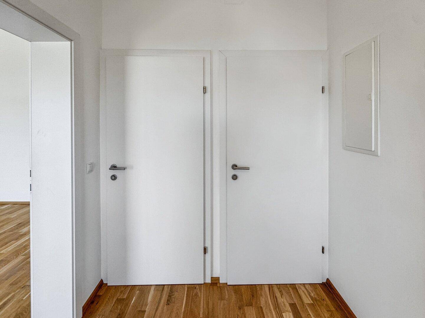 Heller Flur mit zwei weißen Türen und einem Sicherungskasten an der Wand.
