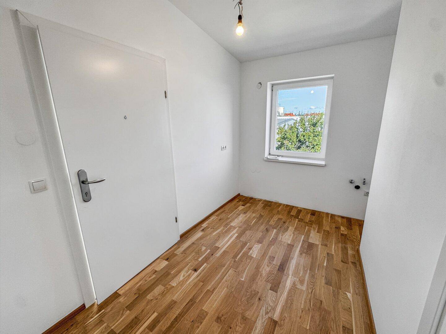 Helles Zimmer mit Parkettboden, weißer Tür und einem Fenster mit Blick ins Grüne.