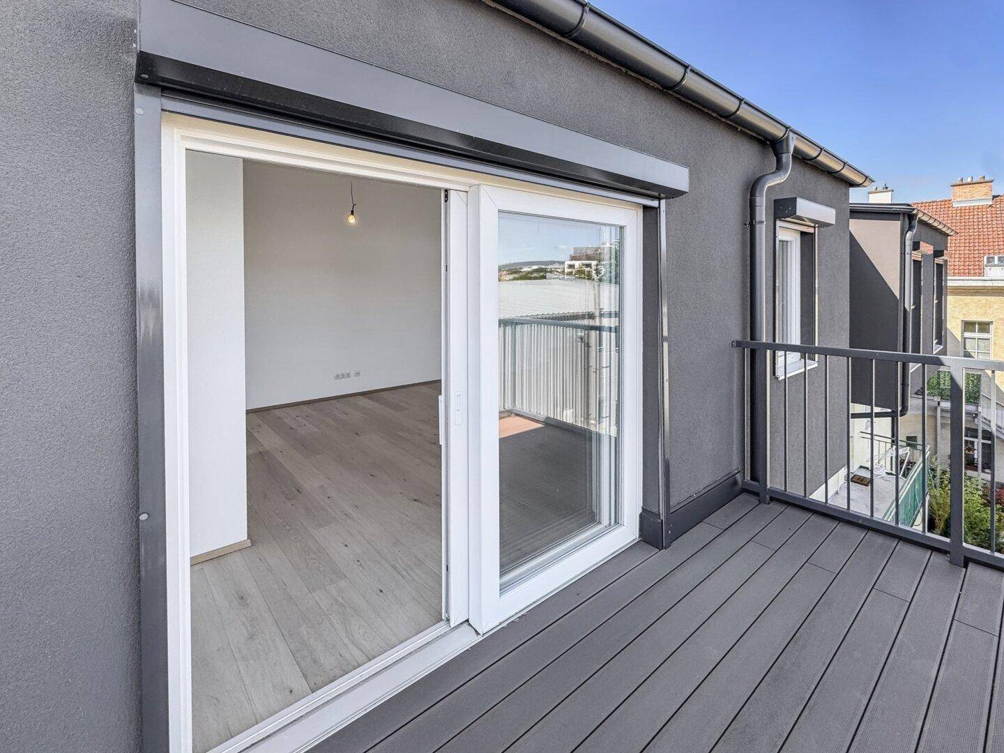 Moderner Balkon mit dunklen Holzdielen und Schiebetür zum Innenraum, ausgestattet mit Rollladen.