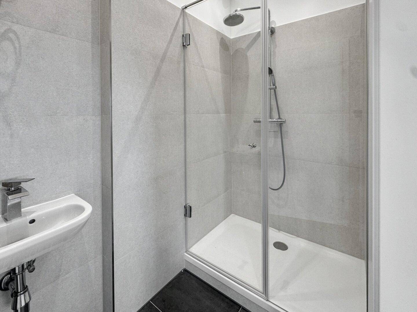 Modernes Badezimmer mit Duschkabine, Regenbrause und kleinem Waschbecken, hell gefliest.