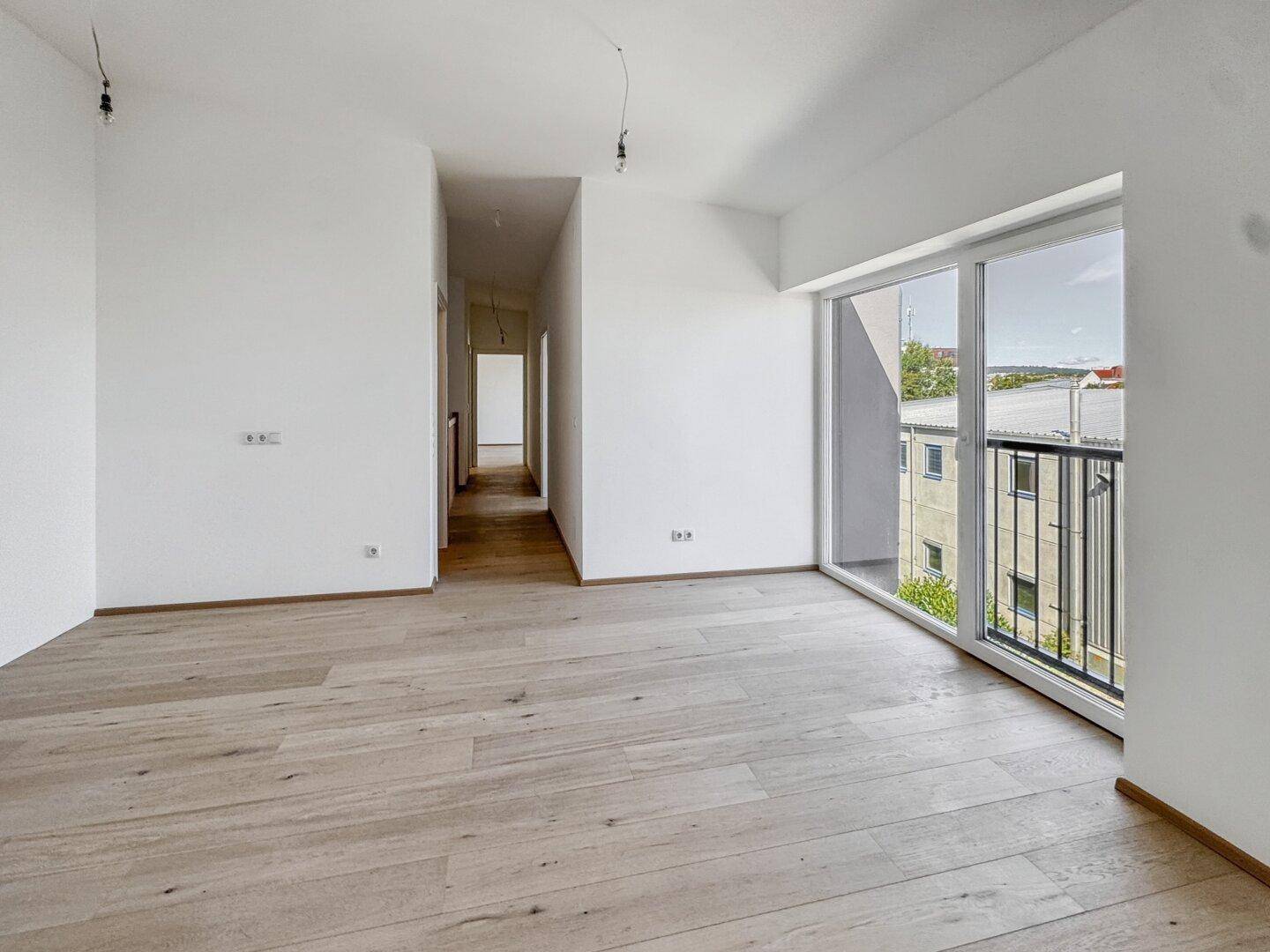 Heller Wohnbereich mit Parkettboden und großem Fenster, das Zugang zu einem Balkon bietet.