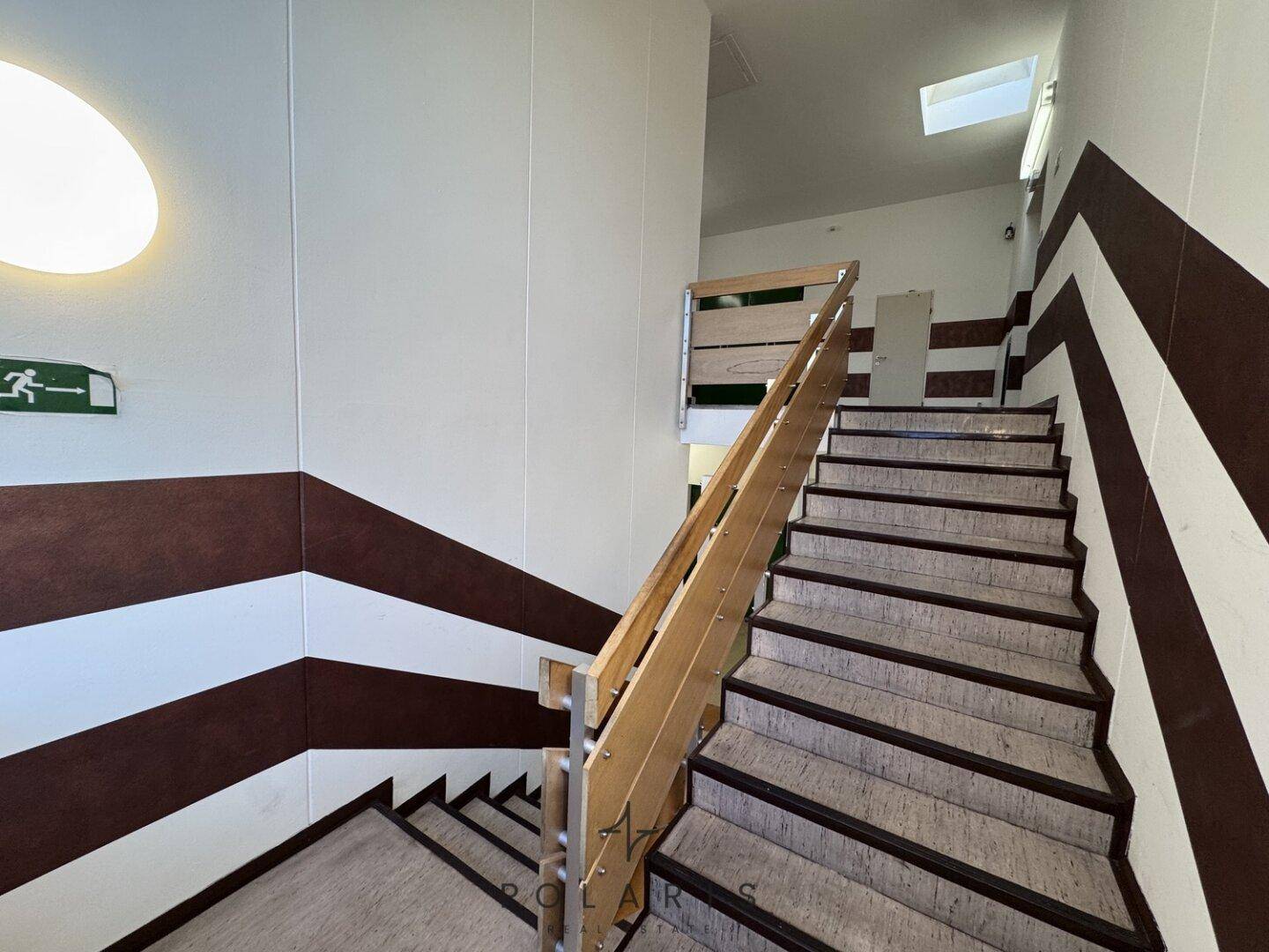 Treppenhaus mit robustem Holzgeländer, gestreiften Wänden und natürlichem Lichteinfall von einem Oberlicht.