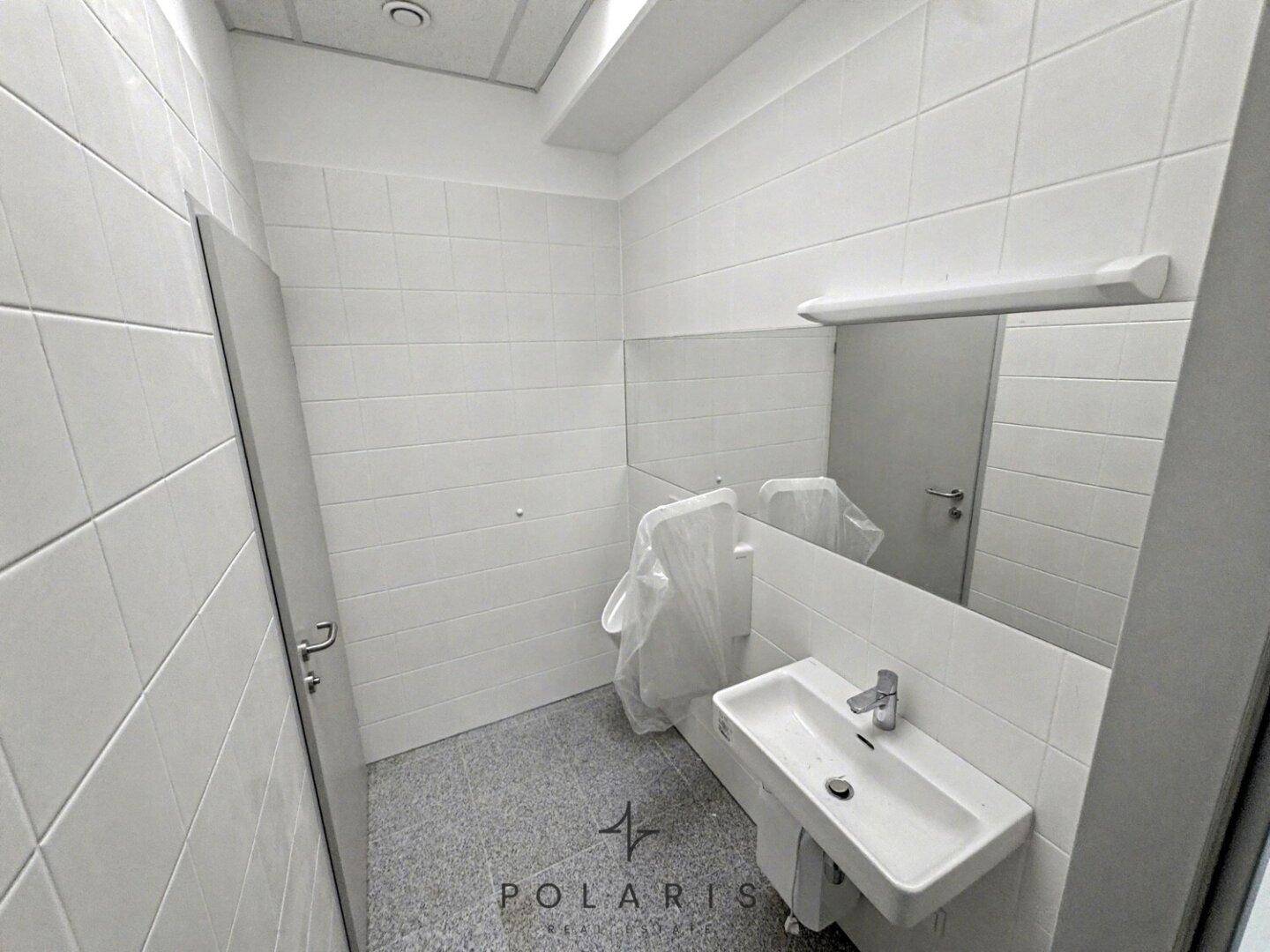 Saubere Toilette mit weißen Fliesen, Waschbecken und Urinal, in gutem Zustand.
