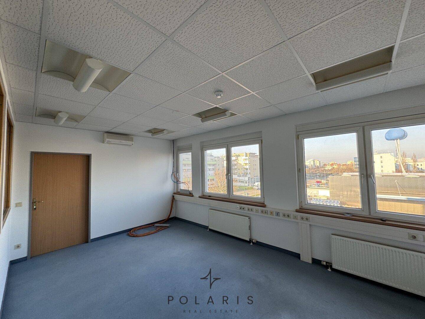 Leeres Büro mit Teppichboden, mehreren Fenstern und einer Klimaanlage an der Wand.
