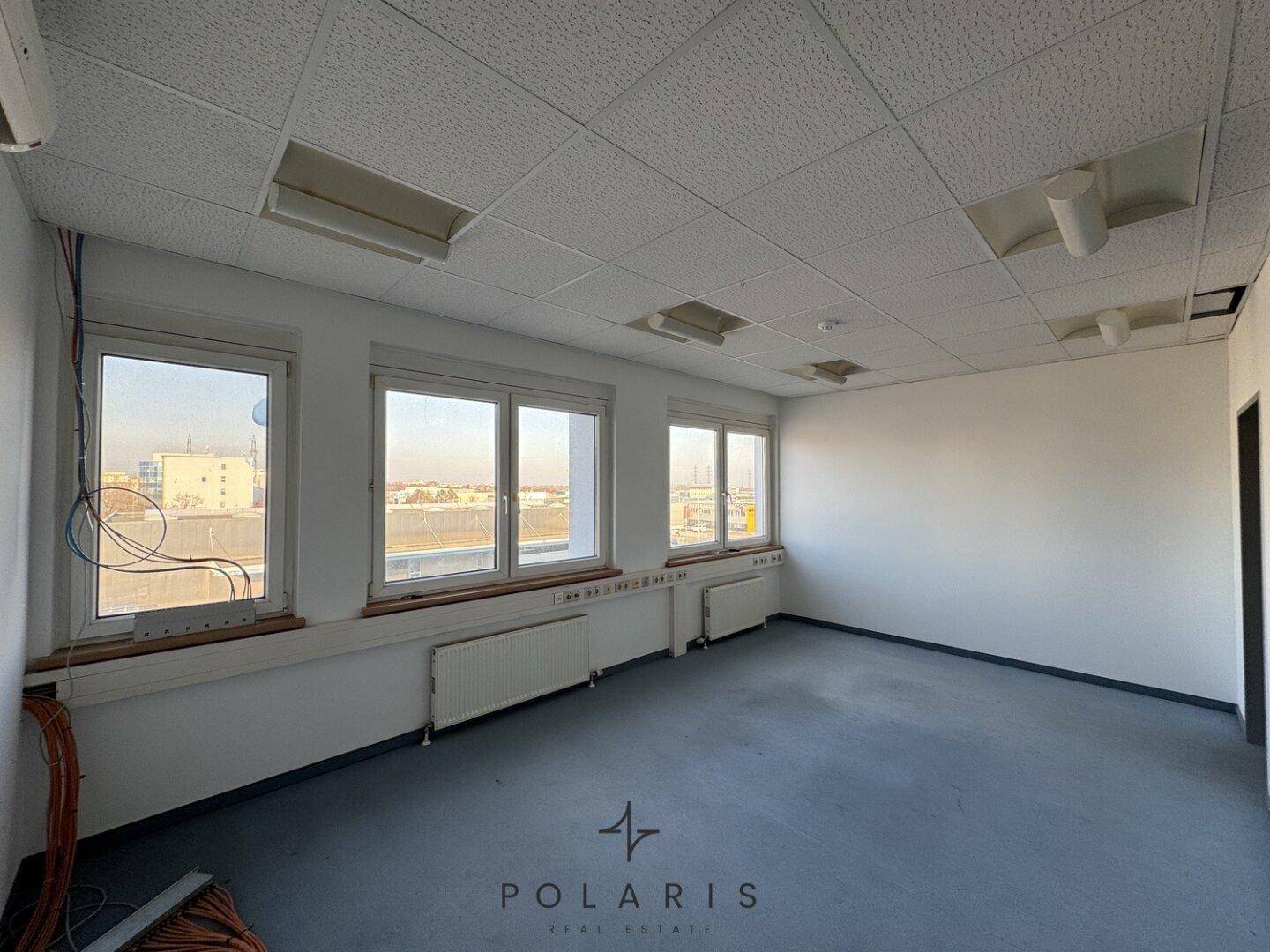 Büro mit Teppichboden, vielen Fenstern und sichtbaren Kabeln entlang der Wand.
