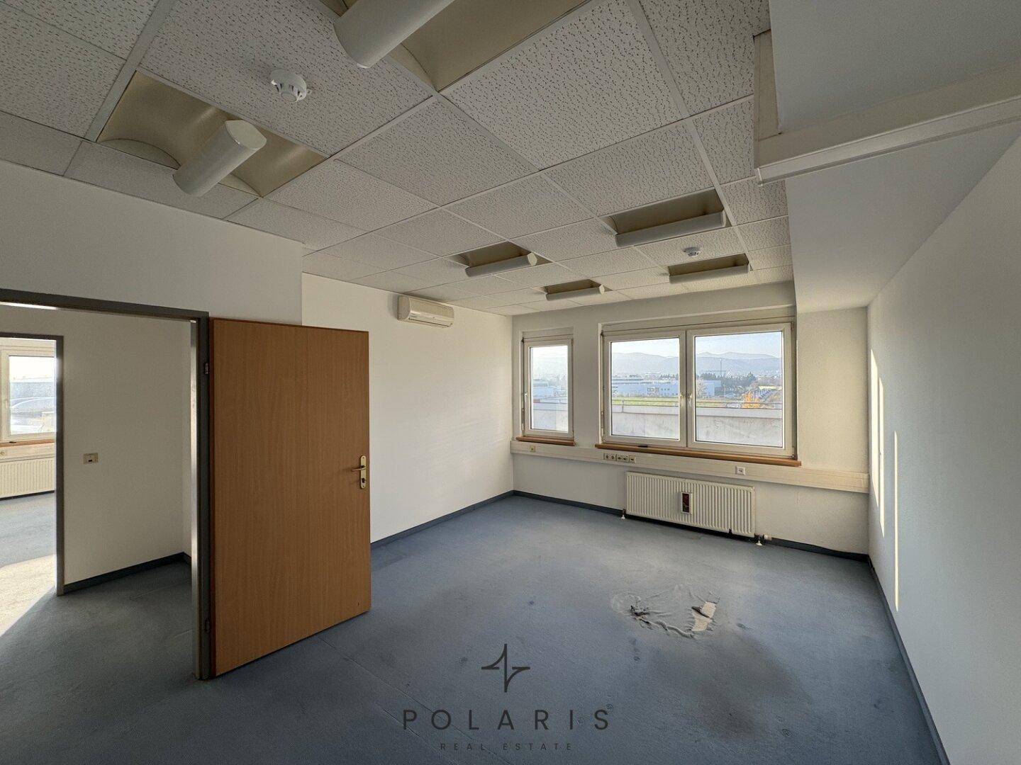 Büro mit Teppichboden, Fenstern und einer Tür, die zu einem weiteren Raum führt.