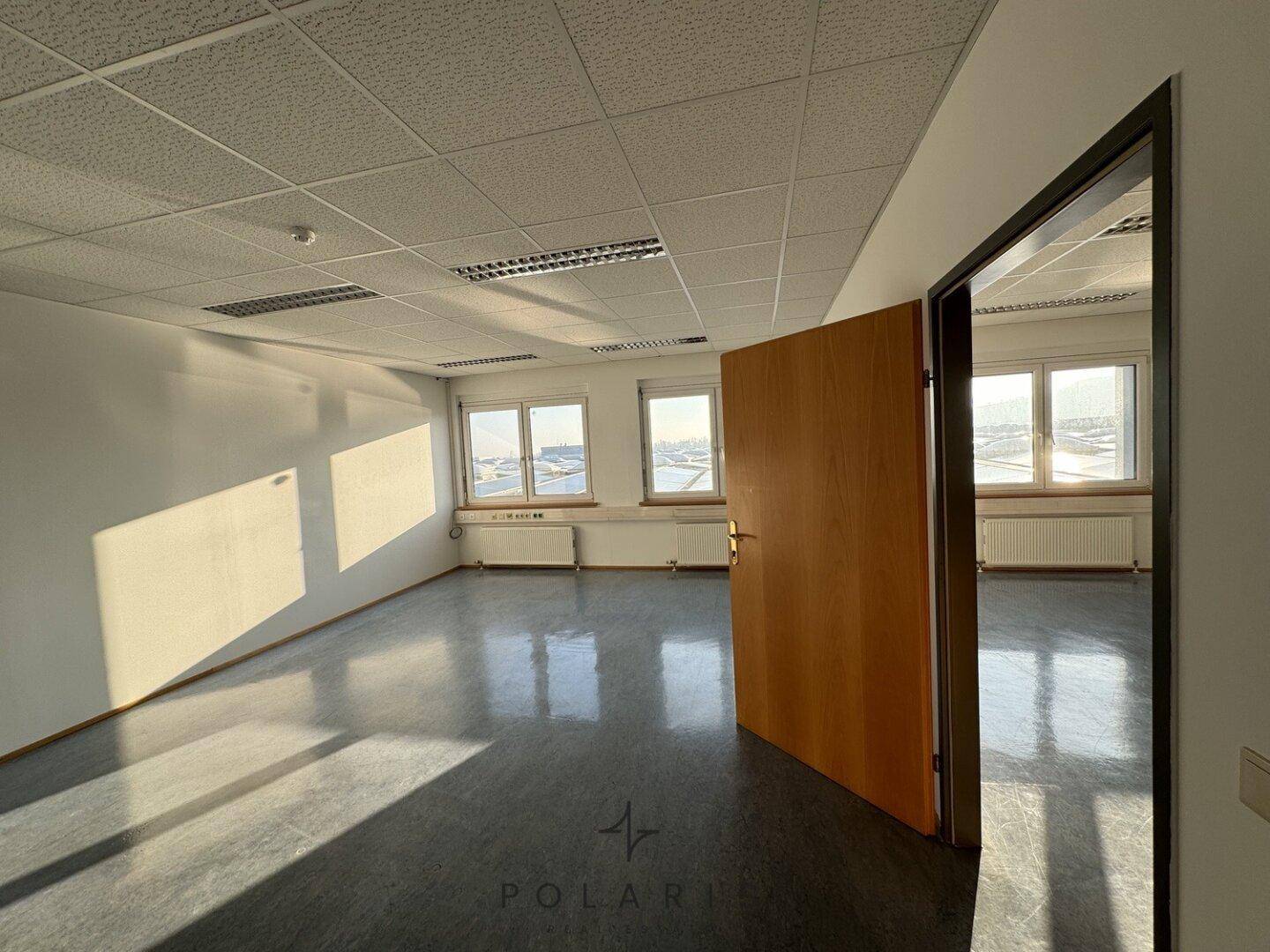 Leeres Büro mit offener Holztür, die den Zugang zu weiteren Räumen ermöglicht, und Vinylboden.