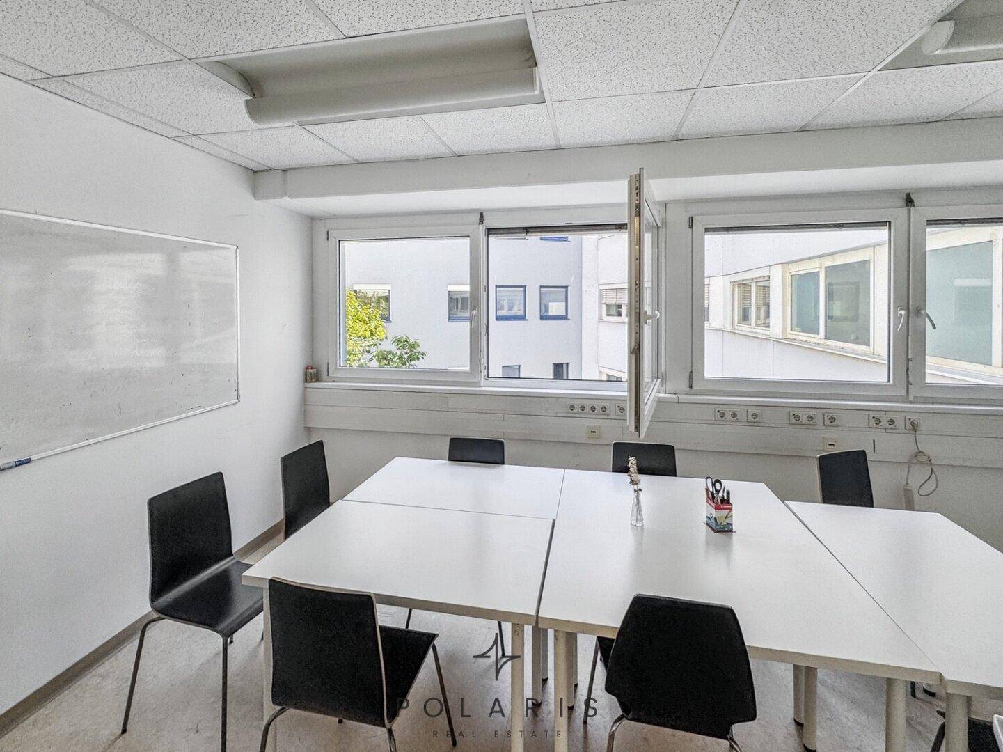 Heller Besprechungsraum mit weißem Tisch, schwarzen Stühlen und einer Whiteboard-Wand, gut beleuchtet.