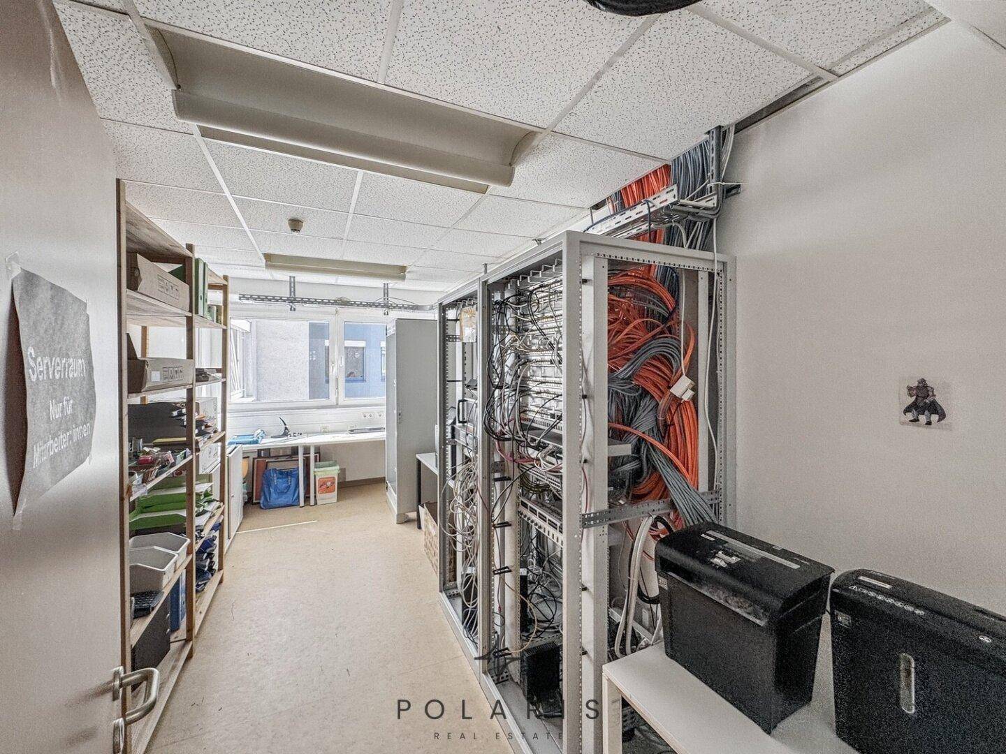 Serverraum mit offenen Racks, Verkabelung und Lagerregalen, beleuchtet durch Deckenleuchten.