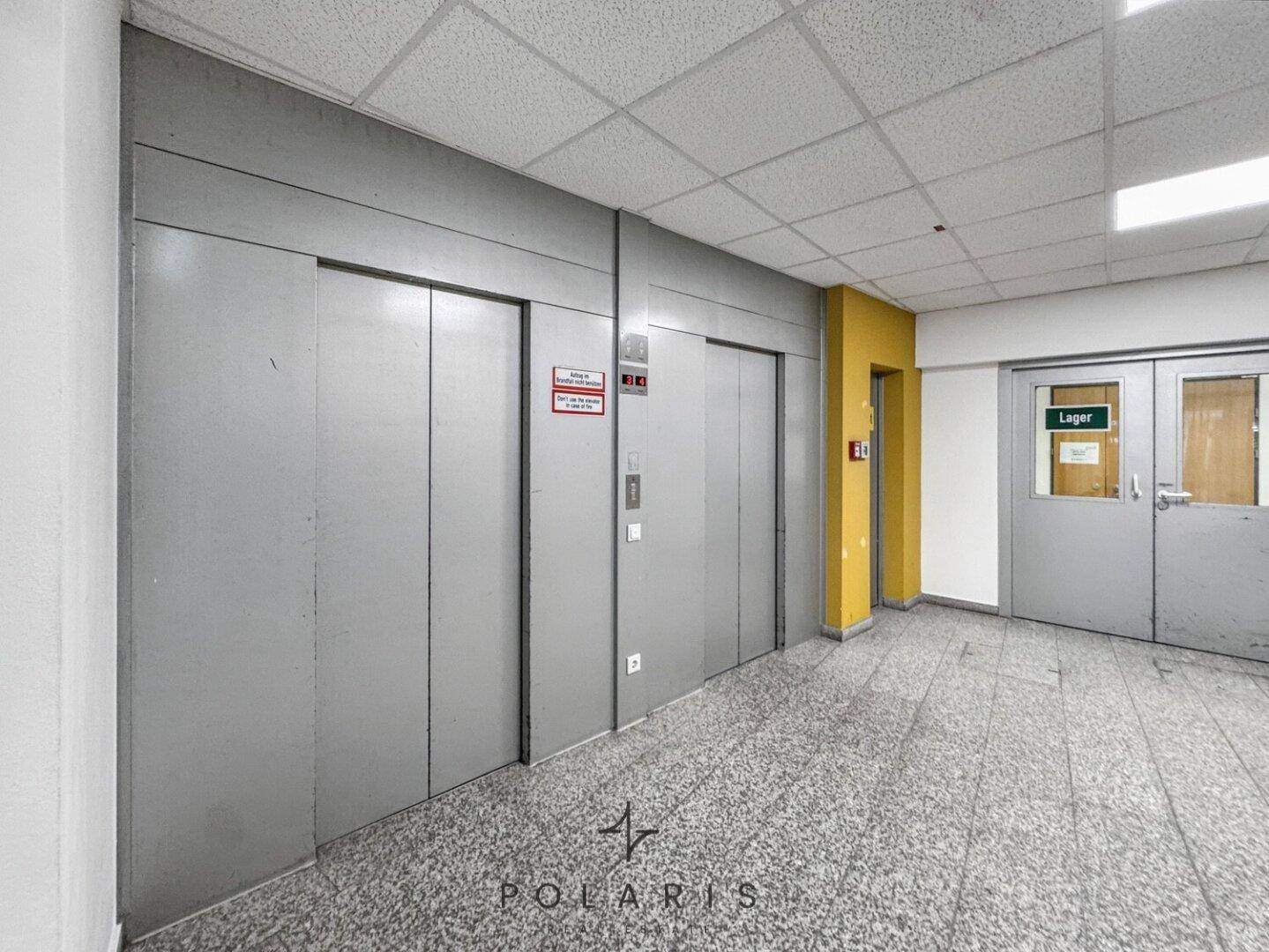 Aufzugsbereich mit zwei Metalltüren und einem Lagerraum, gekennzeichnet durch eine gelbe Wand.