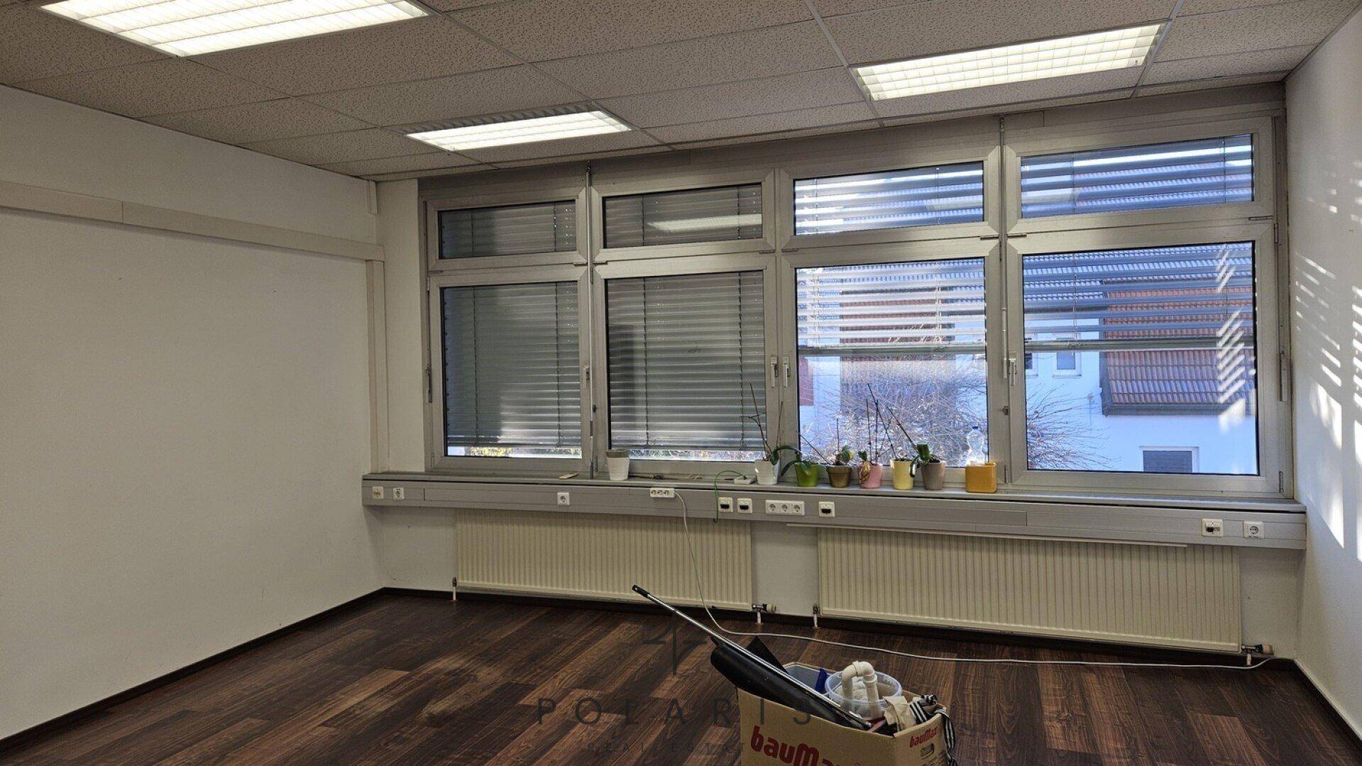 Nahaufnahme der Fensterfront im Büro mit Jalousien, Heizkörpern und Blick auf umliegende Gebäude und Bäume.