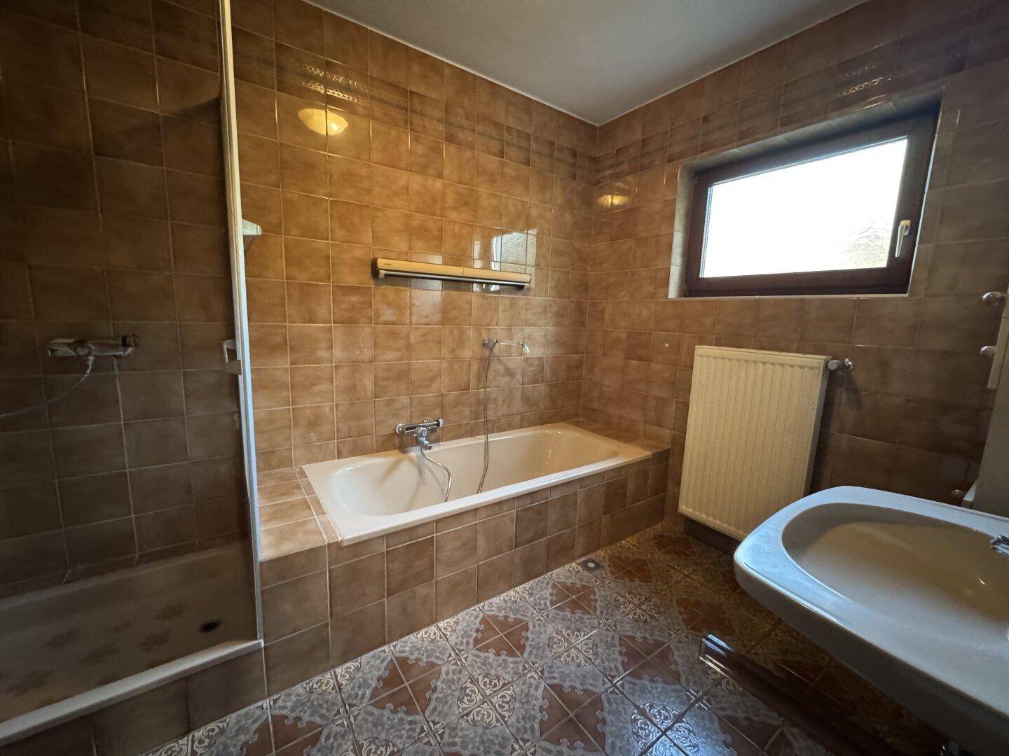 Badezimmer mit Badewanne, Duschbereich, Waschbecken und braunen Fliesen.