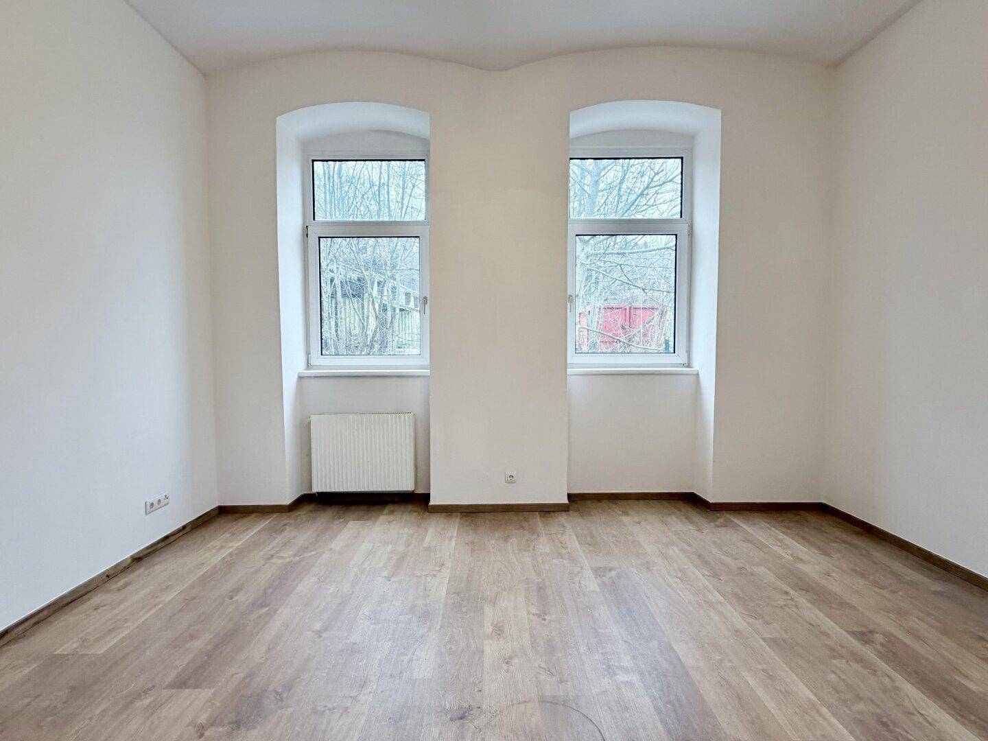 Geräumiger, heller Innenraum mit zwei großen Fenstern und neuwertigem Laminatboden.