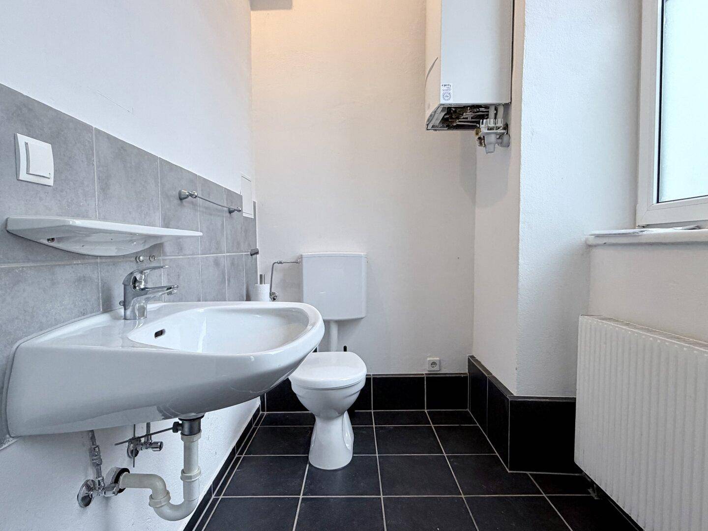 Funktionales Badezimmer mit Waschbecken, Toilette und Gastherme, ausgestattet mit dunklen Fliesen.