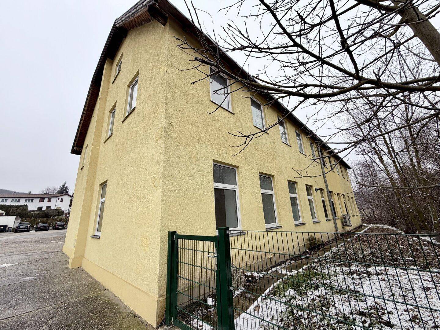 Gelbes Mehrfamilienhaus mit mehreren Fenstern und einem grünen Zaun im Vordergrund.
