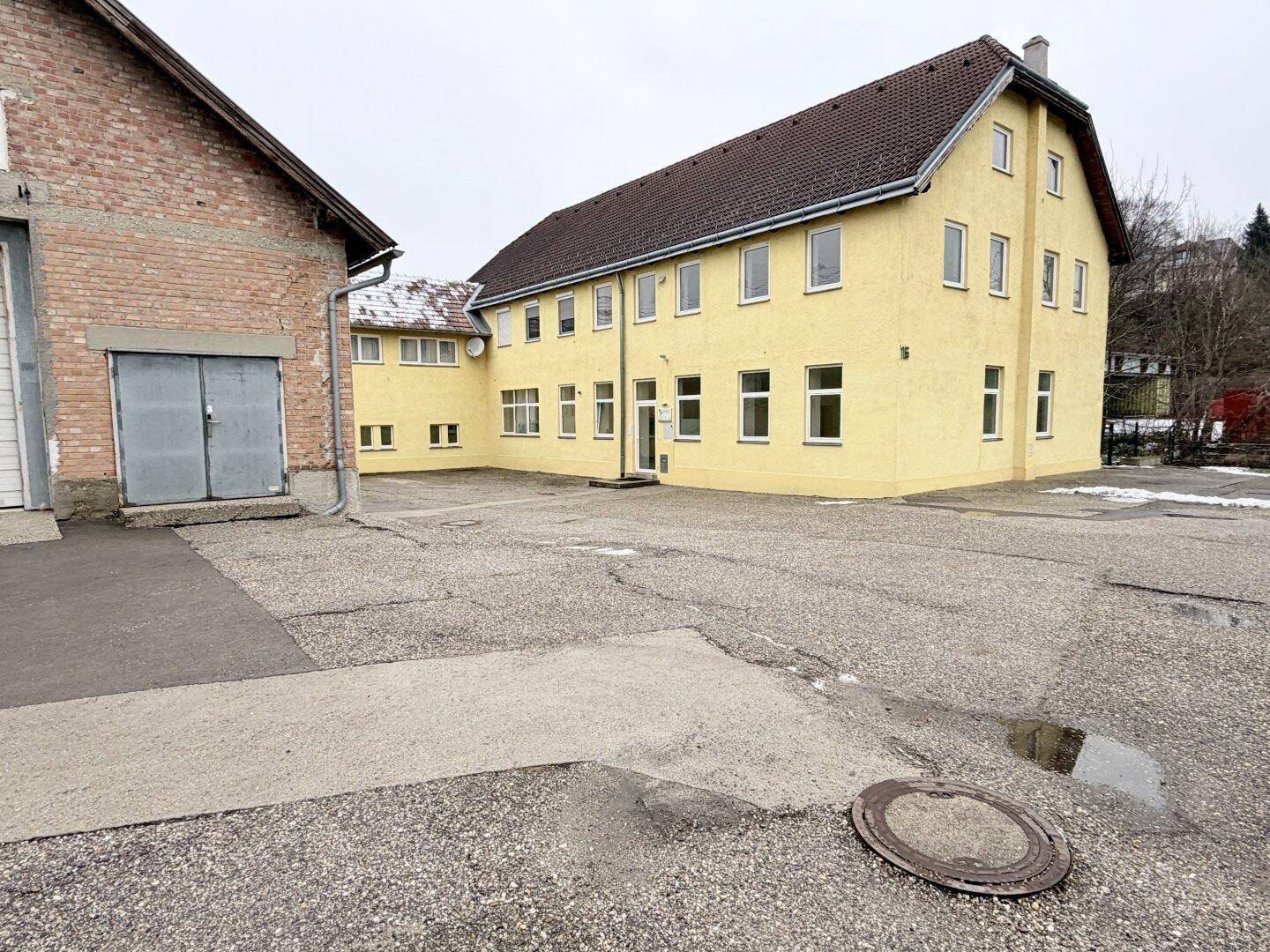 Gelbes Gebäude mit angrenzendem Backsteingebäude und großem Vorplatz, umgeben von Natur.