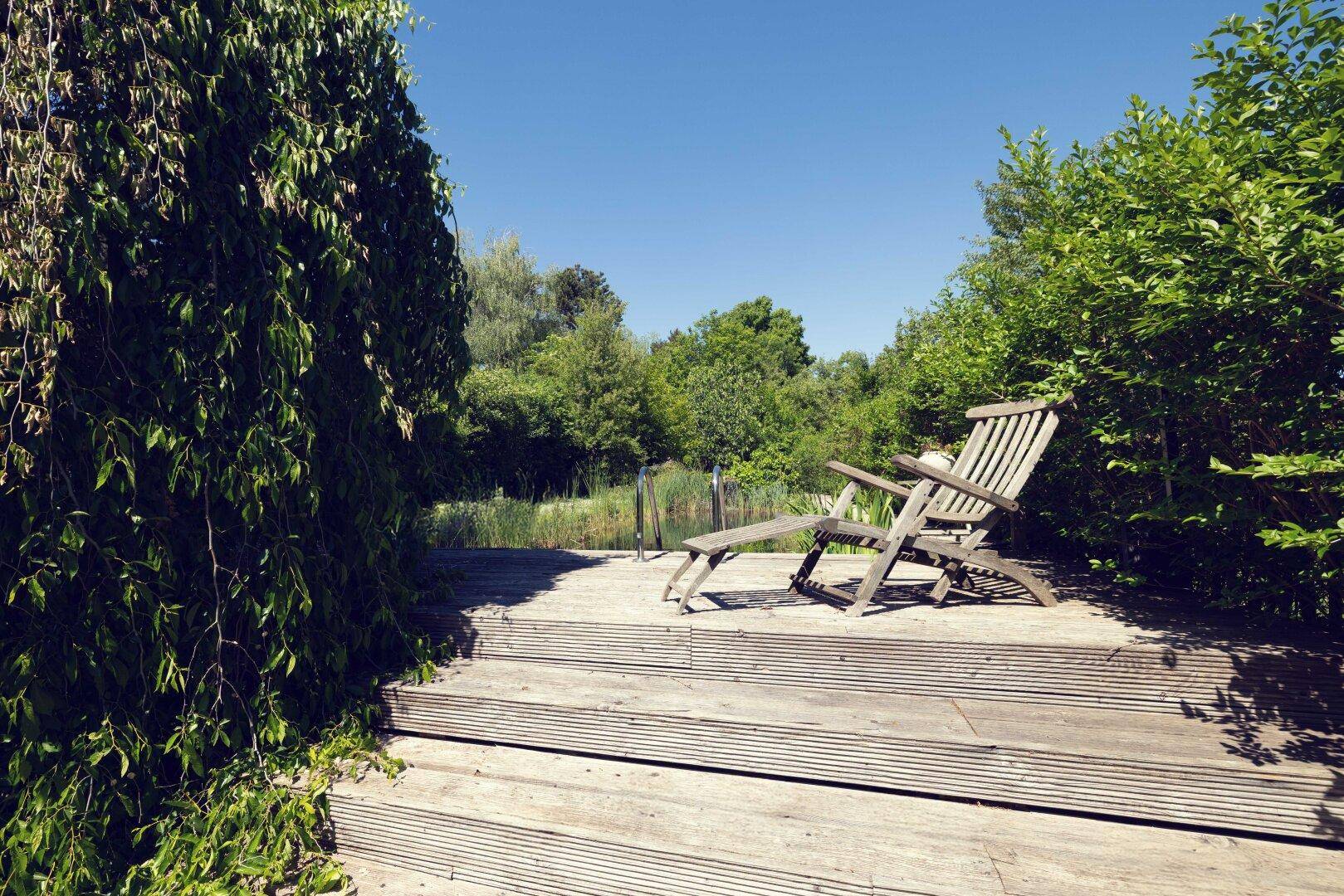Hölzerner Liegestuhl auf einer Holzterrasse am Teich, umgeben von üppiger grüner Vegetation.