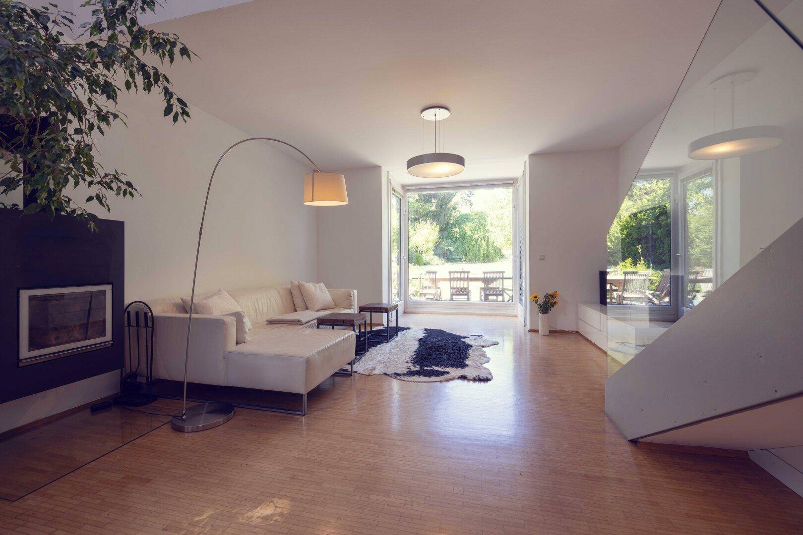 Modernes Wohnzimmer mit weißem Sofa, Kamin und großen Fenstern mit Blick auf den Garten.