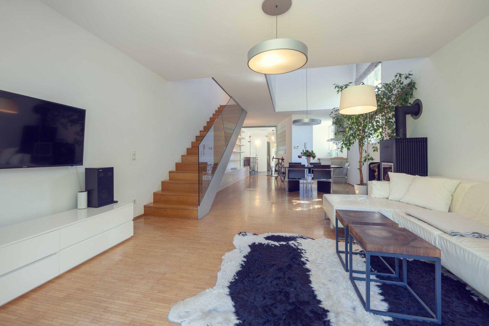 Geräumiger Wohnbereich mit Holzfußboden, weißem Sofa, modernem Ofen und Holztreppe mit Glasgeländer.
