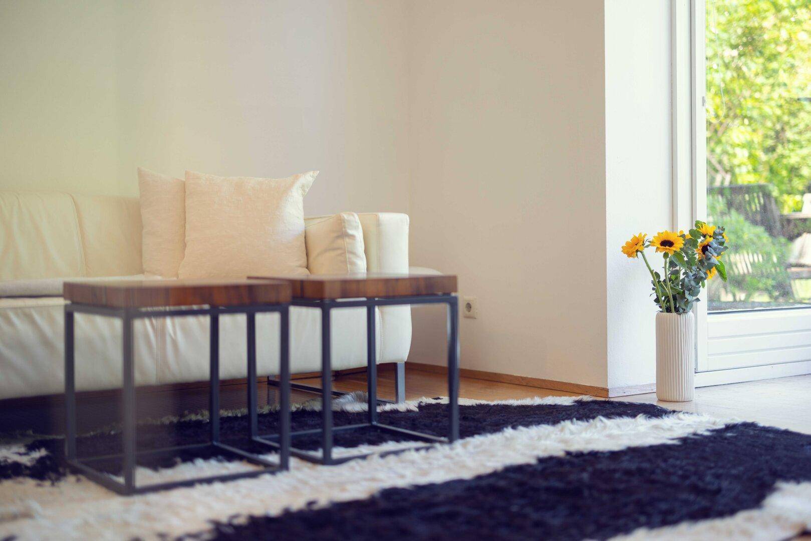 Helles Wohnzimmer mit weißem Sofa, modernen Beistelltischen und Blick ins Grüne durch große Fenster.