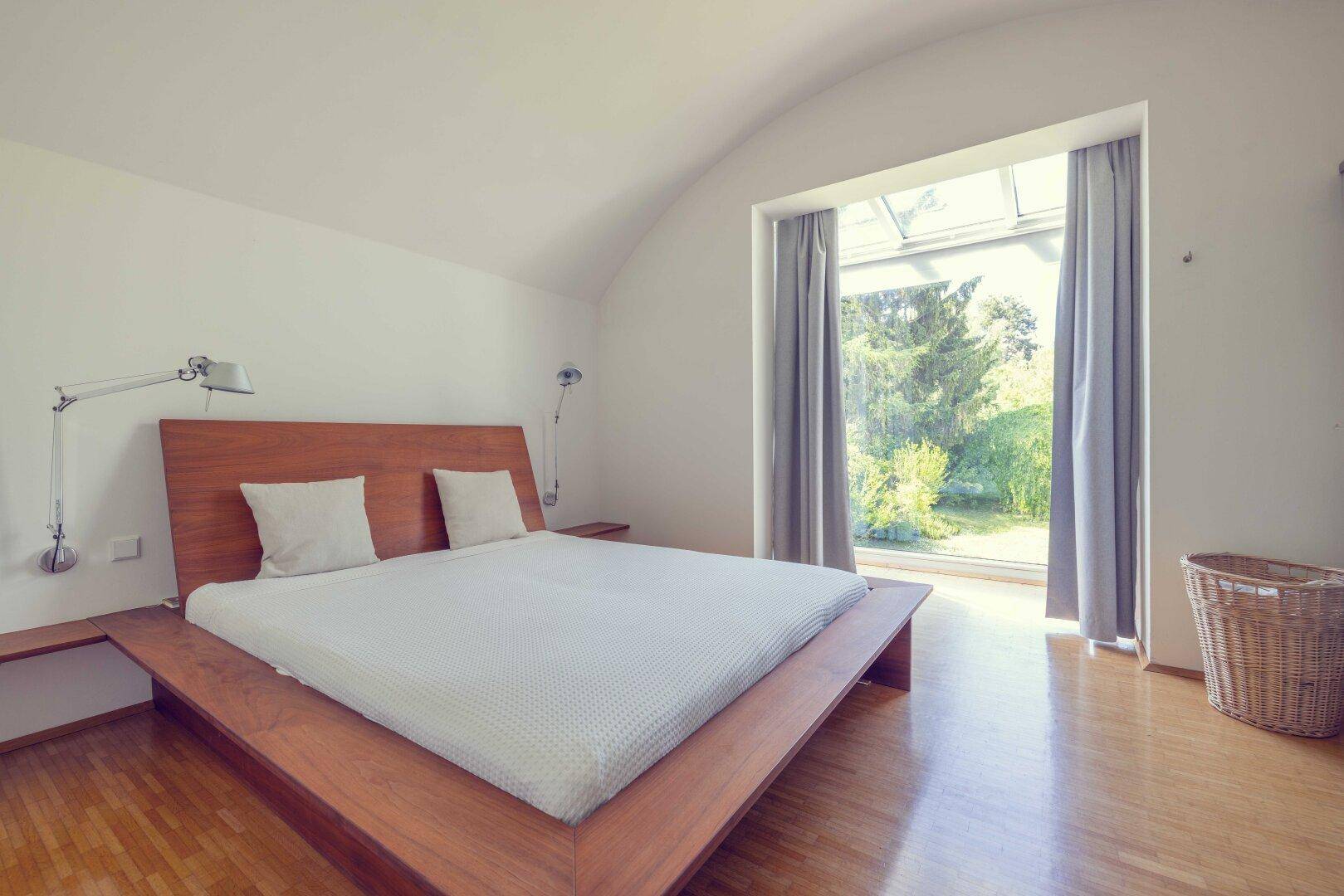 Schlafzimmer mit großem Fenster, Holzbett und Blick ins Grüne, unter einer Gewölbedecke.