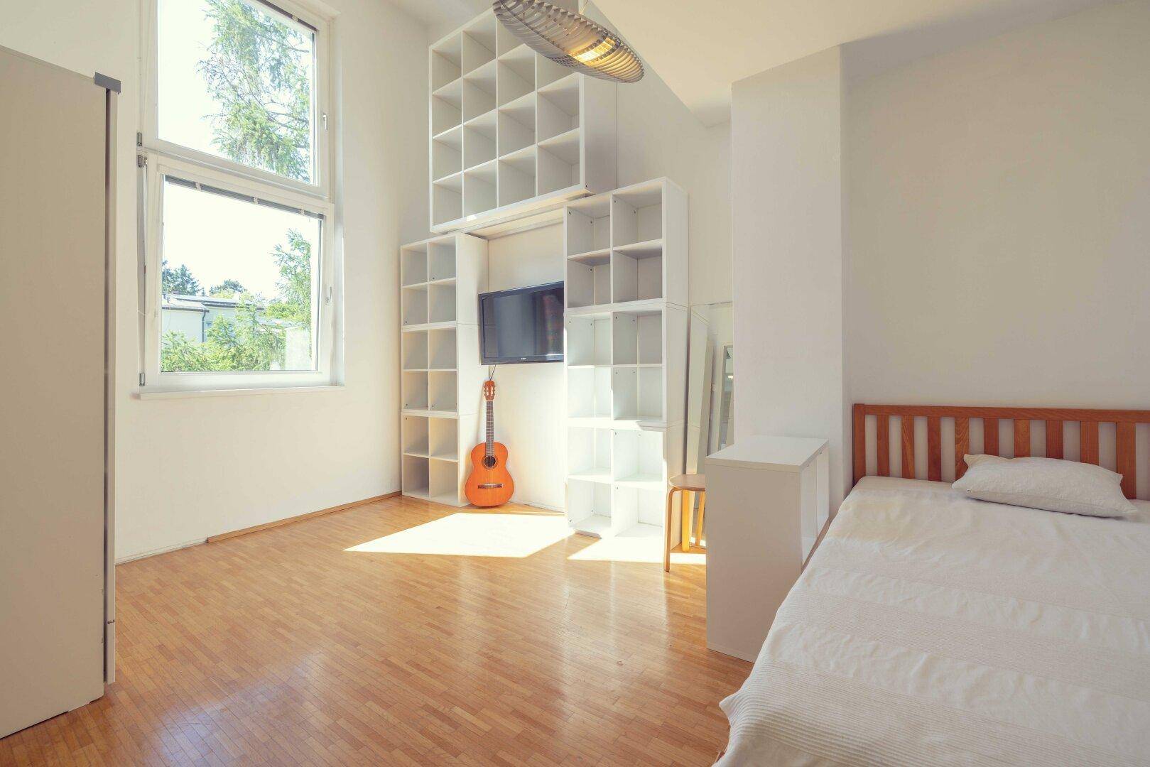 Helles Zimmer mit Holzboden, großem Fenster und modularem Regalsystem mit Fernseher.