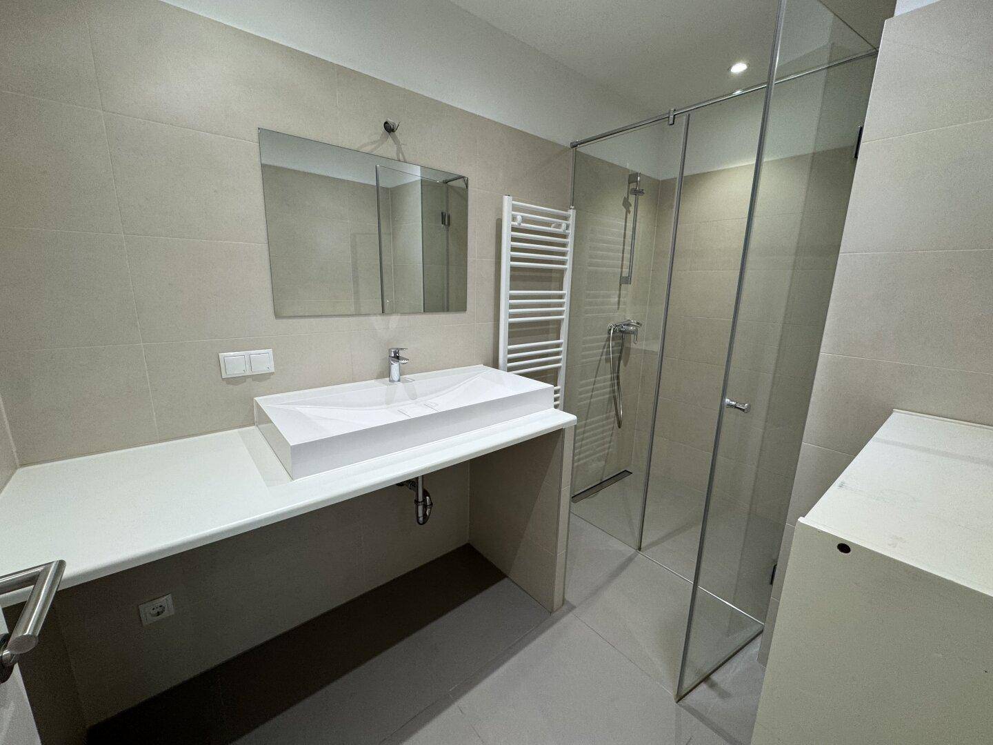 Modernes Badezimmer mit begehbarer Dusche, Einzelwaschbecken, beheiztem Handtuchhalter und hellen Fliesen.