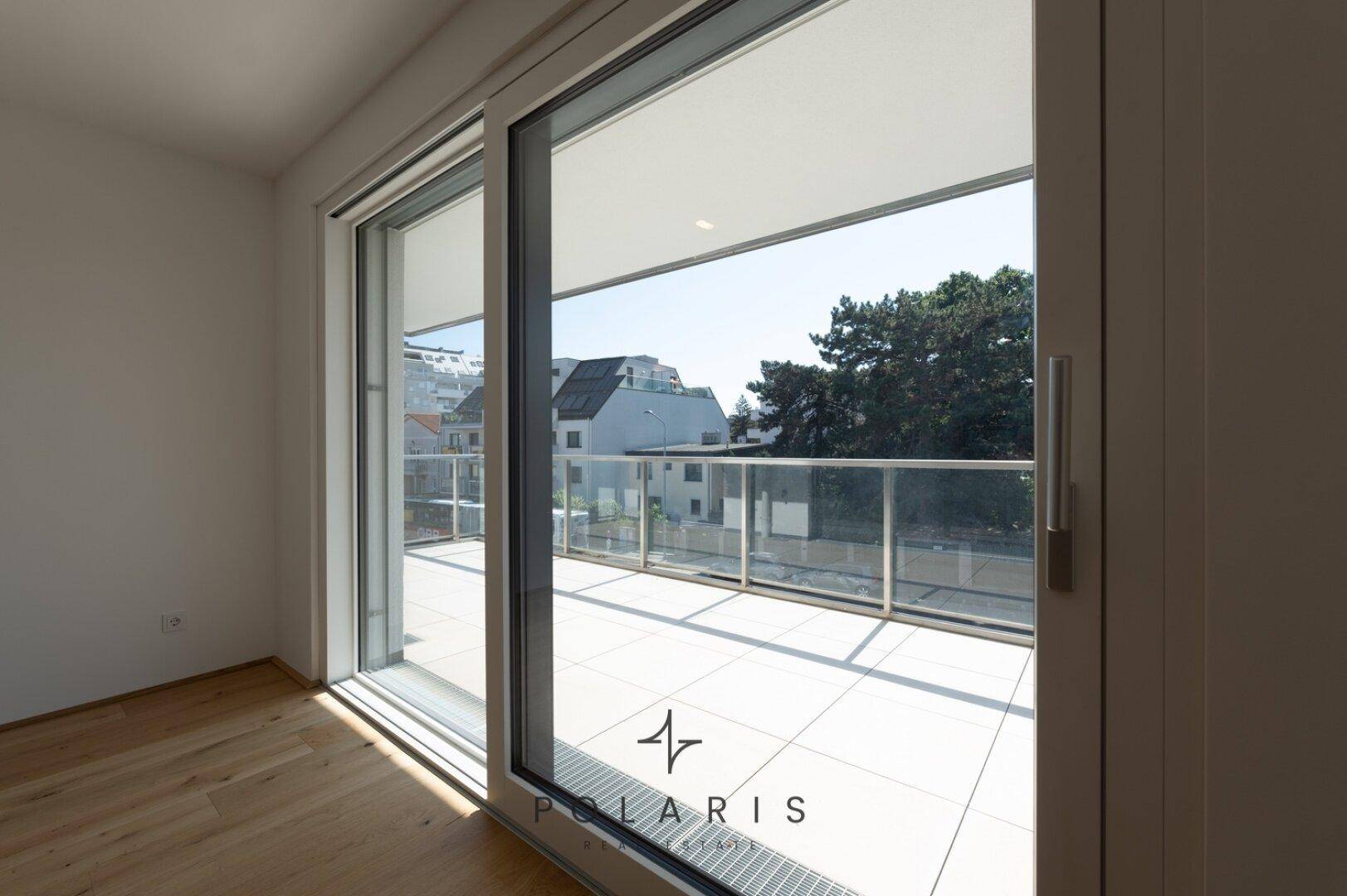 Große Schiebetür führt zu einem sonnigen Balkon mit Glasgeländer und Stadtblick.
